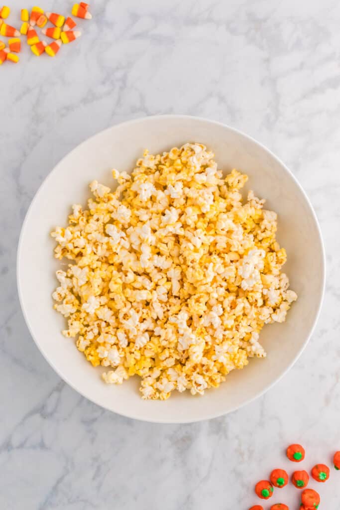 Grand bol blanc rempli de pop-corn fraîchement éclaté sur un comptoir en marbre avec du maïs soufflé et des citrouilles éparpillés autour.